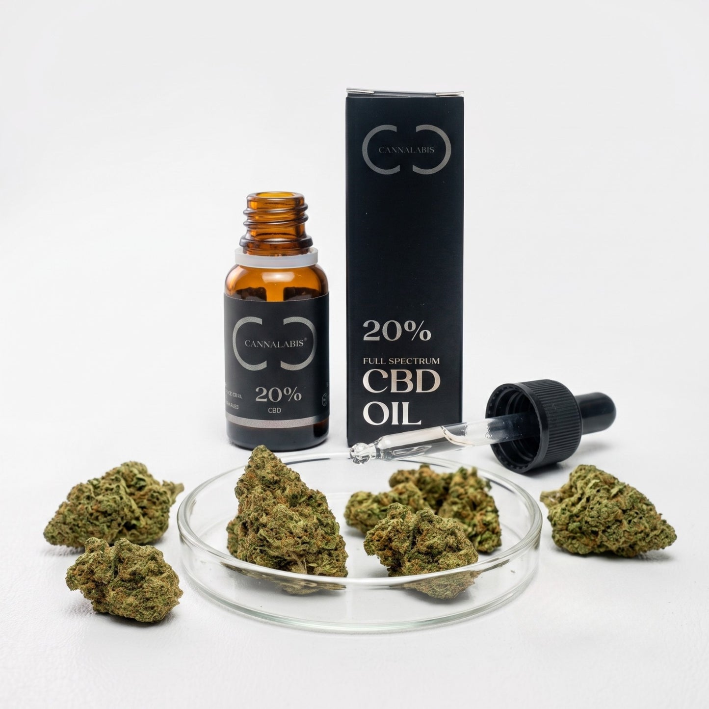 Fotografia de estúdio do Pack Relaxamento sobre um fundo branco. No centro, um frasco de vidro âmbar aberto de óleo de CBD de 20% da marca Cannalabis está de pé ao lado da sua caixa de cartão preta a condizer. O rótulo da caixa indica 'CANNALABIS' e '20% FULL SPECTRUM CBD OIL'. À frente, uma placa de Petri de vidro transparente contém vários brotos secos de cânhamo verde. O conta-gotas de vidro está pousado no bordo da placa, com líquido visível no interior. Outros brotos estão espalhados na superfície.